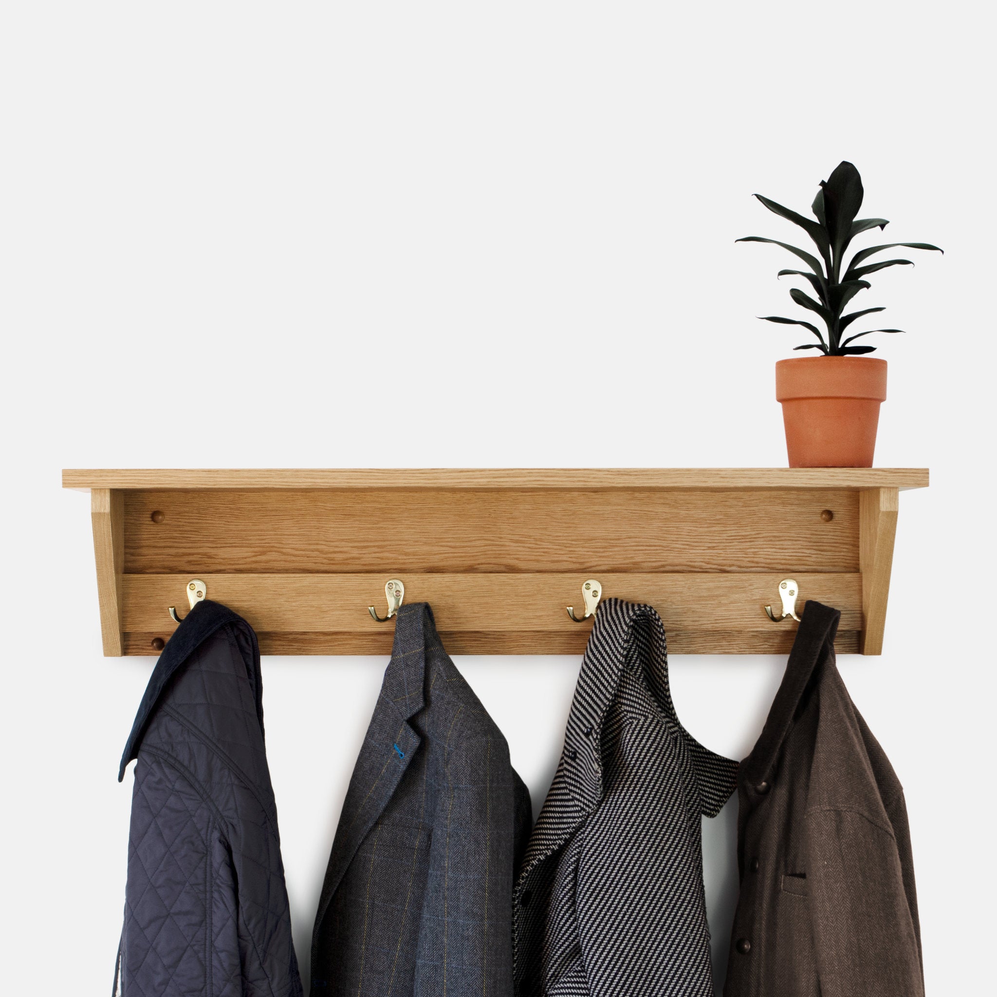 Wooden oak coat rack with hooks holding coats against a white wall, with a potted plant on top.