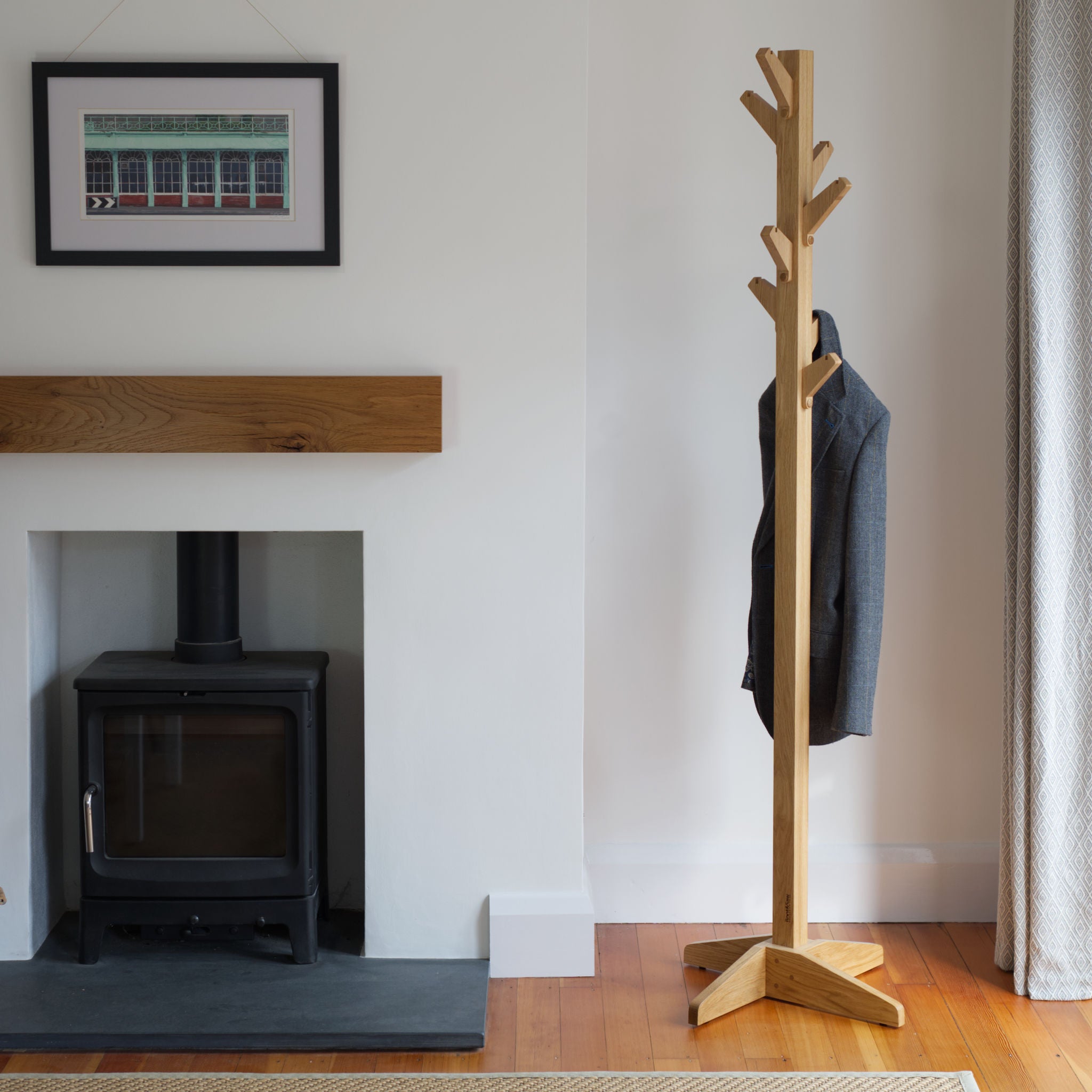 Wooden oak coat tree coat stand with a grey coat against a white wall with a fireplace.